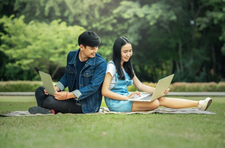 Two students sitting back to back in a park one with long hair and one with short. Each has a laptop but both are looking at the long-haired person's laptop.